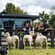 A family of 5 behind a wooden fence at a farm, with 3 sheep and a border collie dog in the foreground in New Zealand